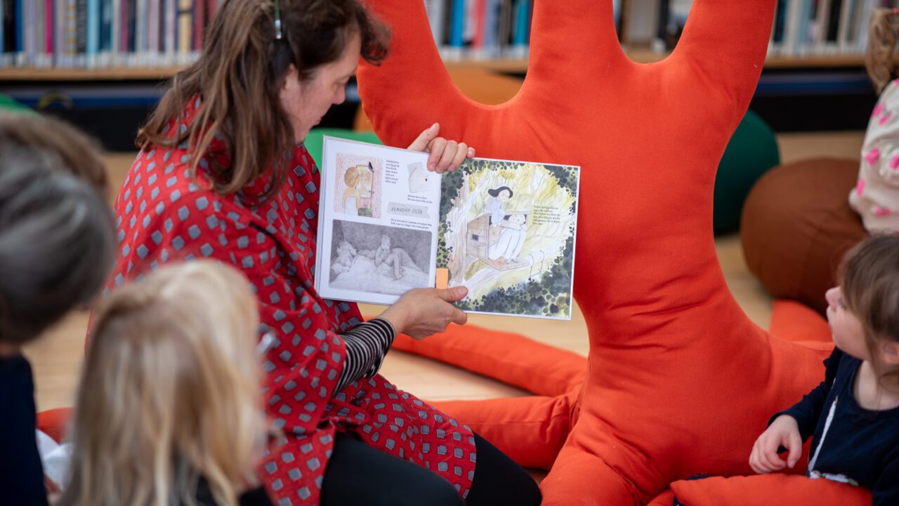 Image of a woman showing the a book to a few kids in the library in the Nordic House
