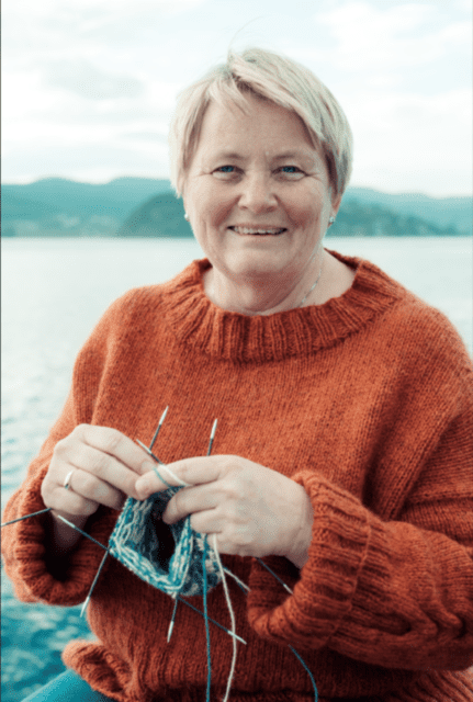 Photo of a woman knitting. She is smiling at the camera, has blond short hair and wearing an orange sweater.