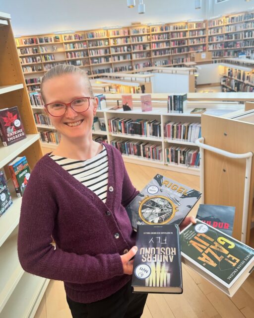 A woman holding 4 books, she is blond and wears glasses and a purple sweater.